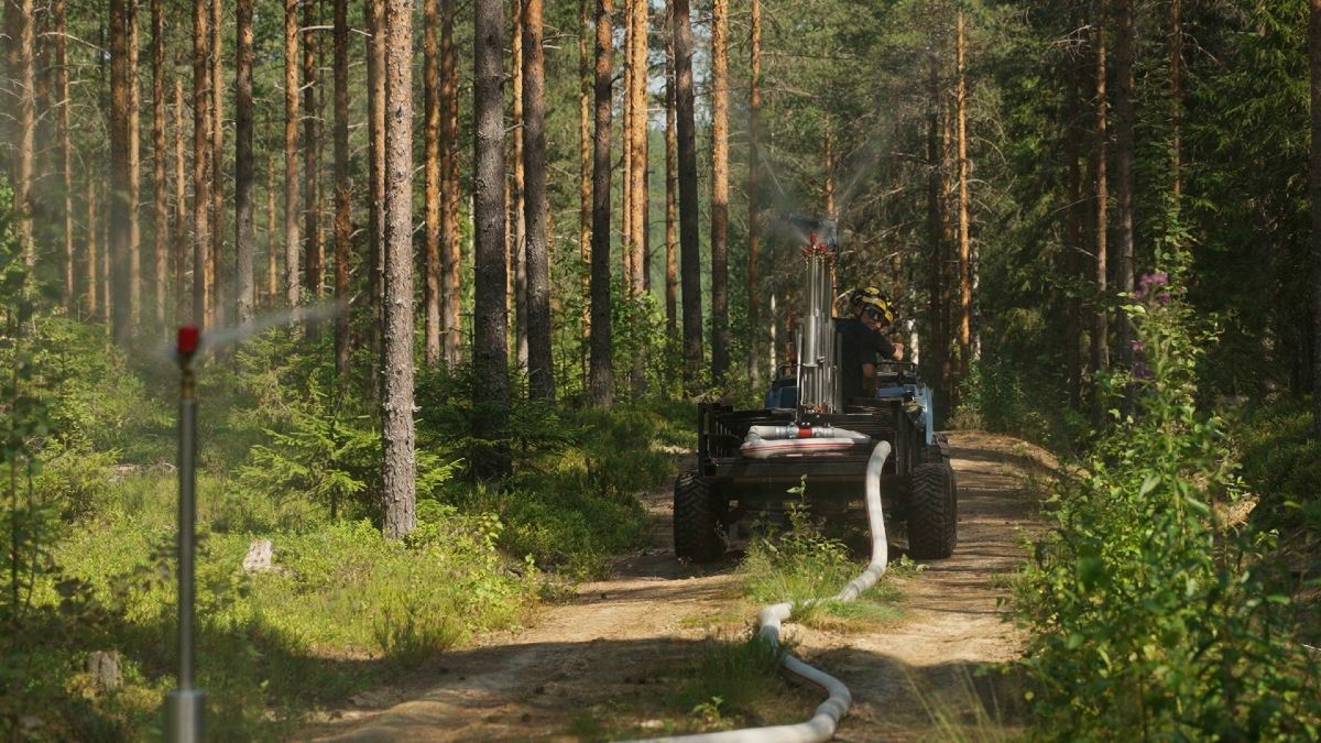 Bilden visar ett skogslandskap med många höga träd. En grusväg går genom skogen. Vid vägkanten finns en slang och en bevattningsanordning. I slutet av vägen syns ett fordon med en person och utrustning på. En slang går från fordonet längs grusvägen.