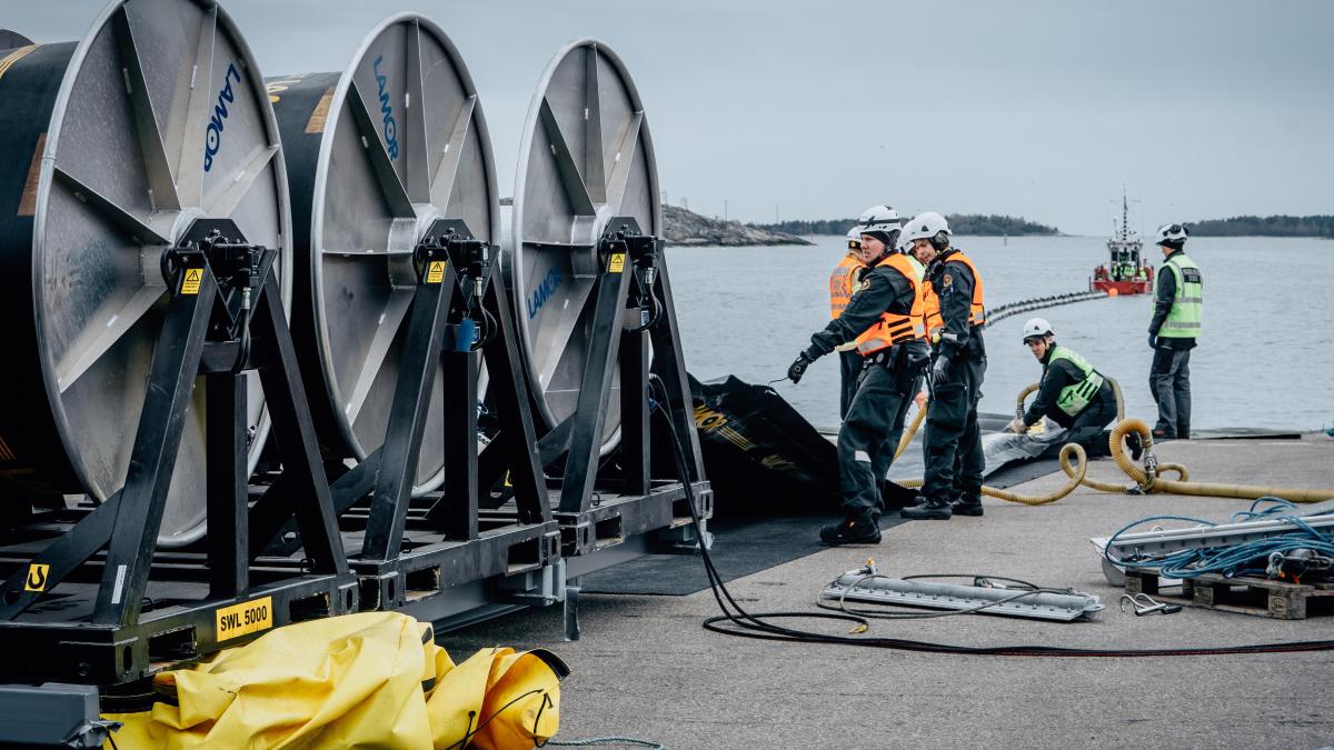 Oljeskyddspersonal arbetar på ett hamnområde bland stora kabeltrummor och bekämpningsutrustning vid havet.