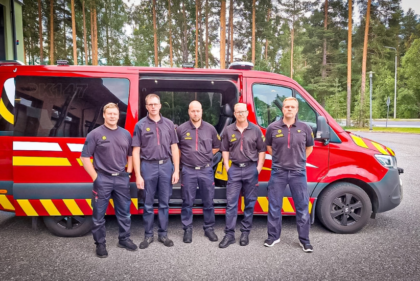 Viisi henkilöä seisoo hälytysajoneuvon edessä aseman pihalla.