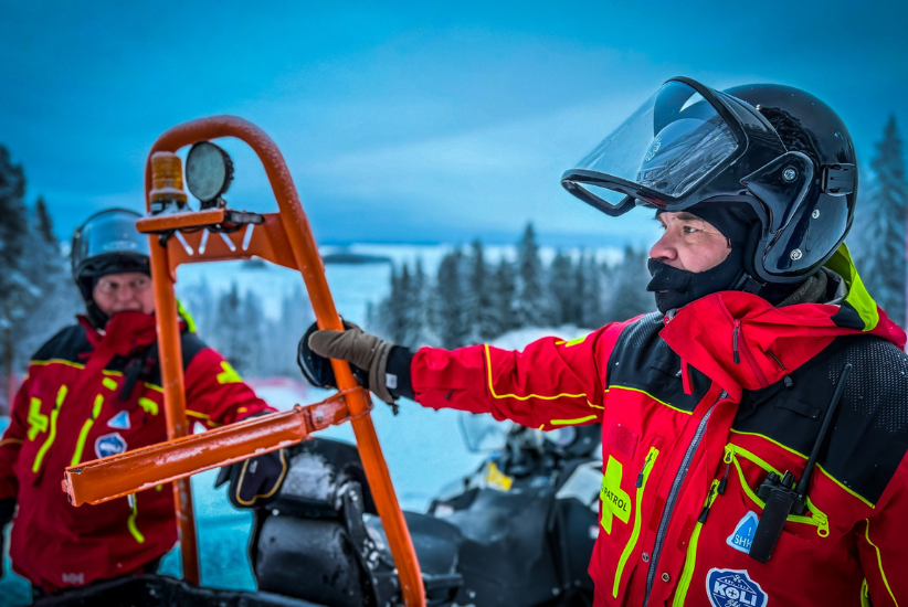 Kaksi henkilöä seisoo lasketturinteellä moottorikelkan vieressä.