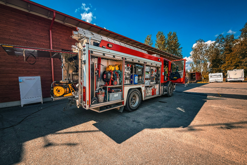 Hälytysajoneuvo, jossa tavaratila sivusta on auki. Taustalla pelastusasema.