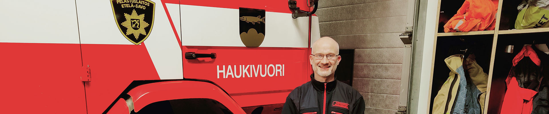 Haukivuoren sopimuspalokuntalainen Mika Karppinen seisoo paloauton ja sopimuspalokuntalaisten varustekaapin vierellä Haukivuoren paloaseman kalustohallissa.