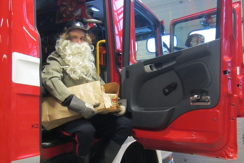 Joulupukiksi pukeutunut henkilö istuu paloauton matkustamossa joulupaketteja sylissään.