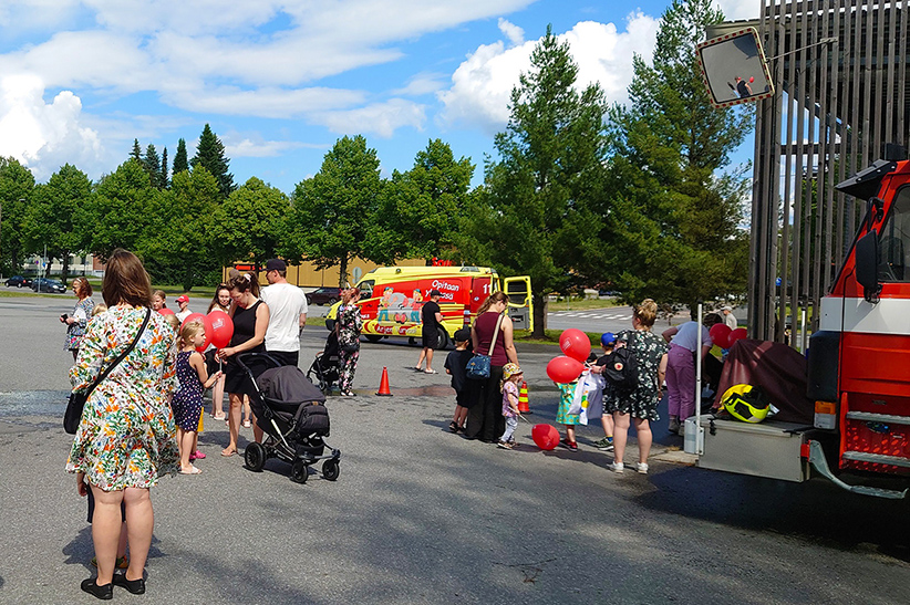 Kuvituskuva pelastuslaitoksen kesätapahtumasta. Kuvassa on aikuisia ja lapsia Mikkelin paloaseman pihalla. Ihmiset ovat kesäisissä vaatteissa, osalla on ilmapalloja.