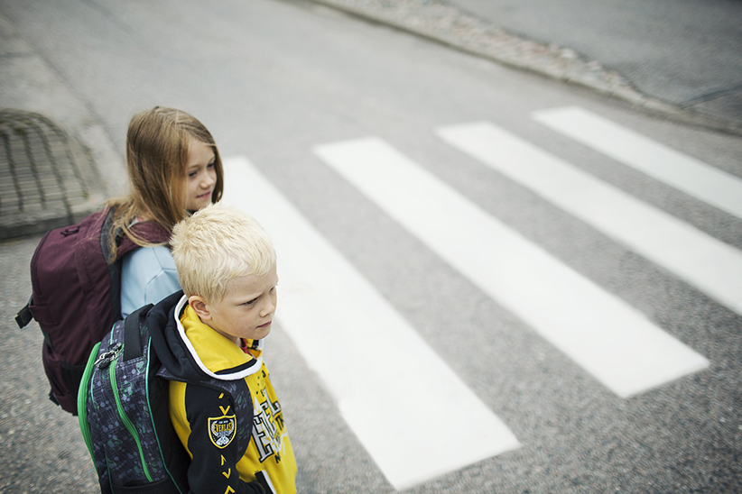 Kaksi pientä koululaista reput selässä menossa suojatielle. Toisella on kirkkaankeltainen paita.