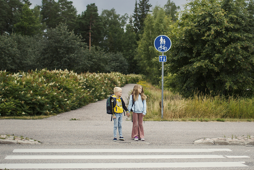 Kaksi koululaista astumassa suojatielle käsi kädessä ja reput selässä.