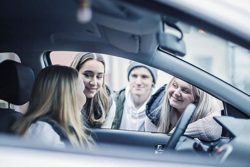 Kuvituskuva: nuori istuu pysäköidyssä autossa kuskin paikalla ja juttelee ikkunasta auton vierellä olevien toisten nuorten kanssa.