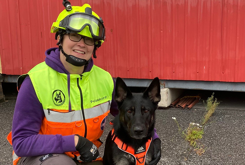 Reetta Koponen pelastuskoiran ohjaajan varusteissaan, huomioliivissä ja kypärä päässä, vierellään saksanpaimenkoira Muru.