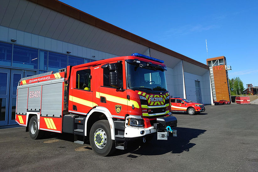 Pelastuslaitoksen kalustoa (sammutusauto ja henkilöstönkuljetusauto) Savonlinnan paloaseman edustalla. Taustalla näkyy torni, jonka seinällä on numero 112.
