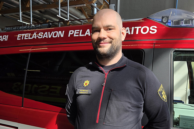 Tomi Raittila katsoo suoraan hymyillen ja nojailee palomestarin autoon.