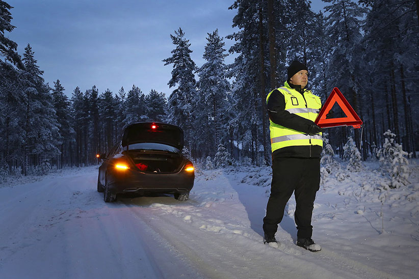 Kuvituskuva: Auto on jäänyt tienposkeen hämärällä ja lumisella tiellä. Kuljettaja on viemässä heijastinliivi päällä varoituskolmiota tien reunaan varoitukseksi takaa tuleville autoilijoille.