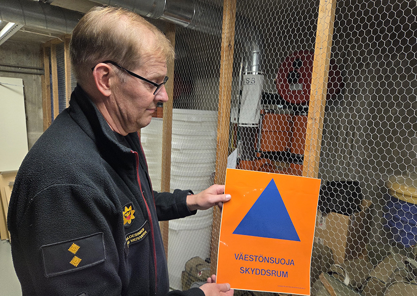 Paloesimies Tomi Merivirta esittelee väestönsuojan merkkiä. Taustalla näkyy väestönsuojan varusteita.