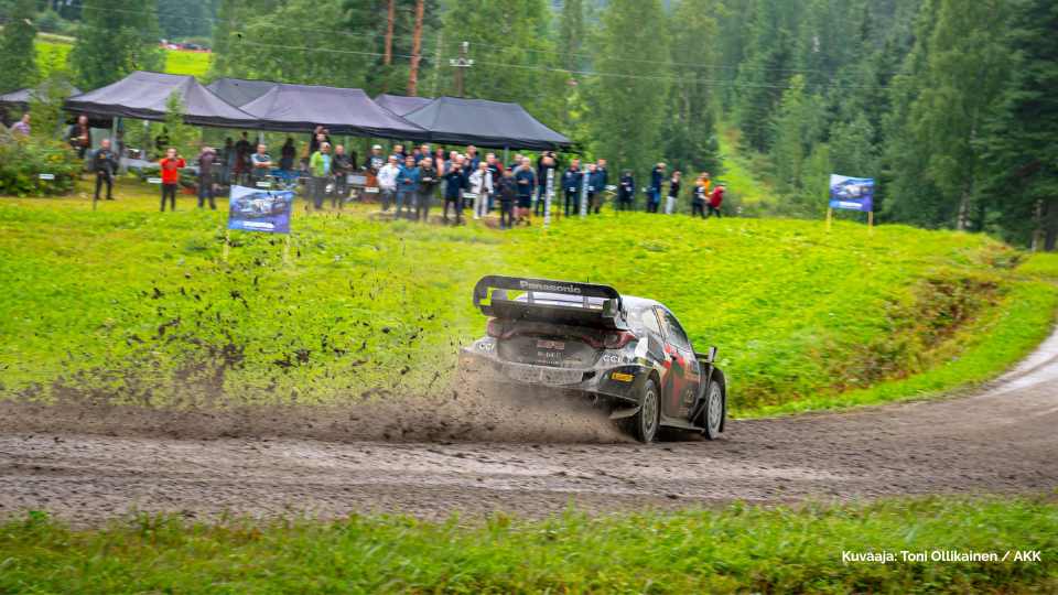 Ralliauto ajaa hiekkatiellä ja kauempana on yleisöä katselemassa