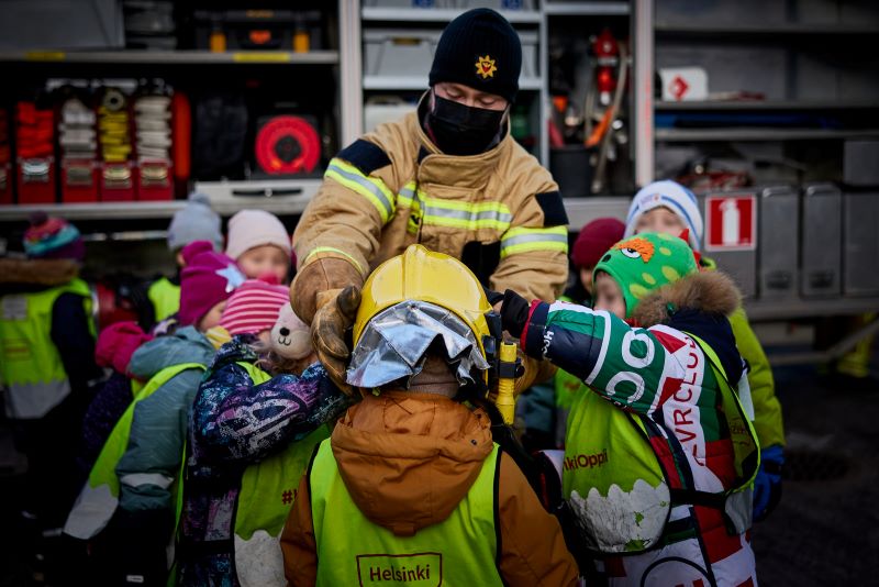 Palomies auttaa lapsia sovittamaan palomiehen kypärää.