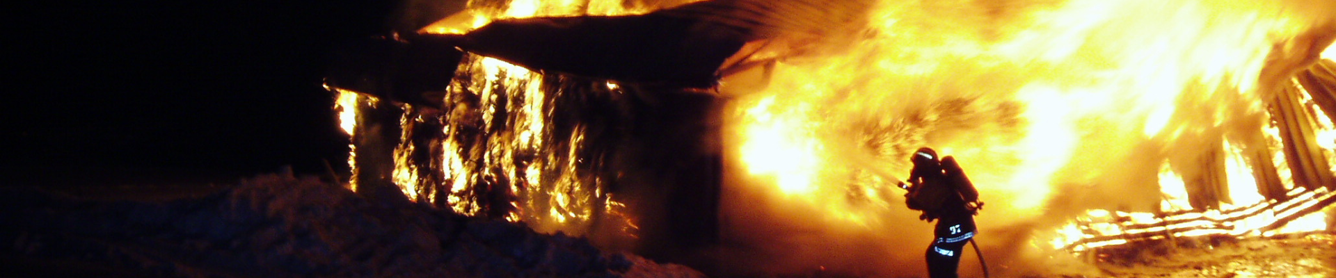 Pelastaja sammuttaa isoa tulipaloa. Vain pelastajan heijastimet ja siluetti erottuvat suurien lieskojen edessä.