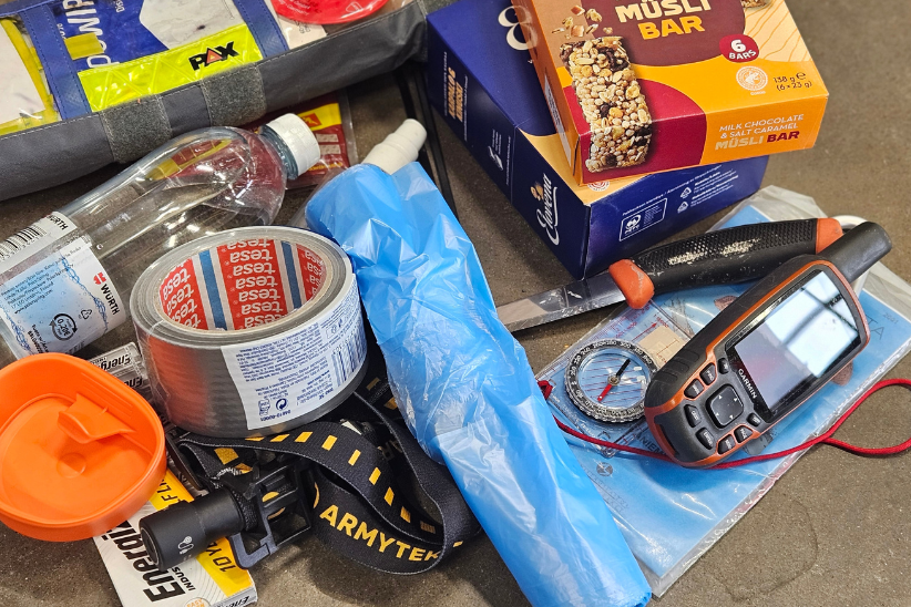 Retkeilijän varautumisvälineistöä kuten ilmastointiteippiä, tulentekovälineet, kompassi, kartta, ruokaa, juomaa ja otsalamppu.