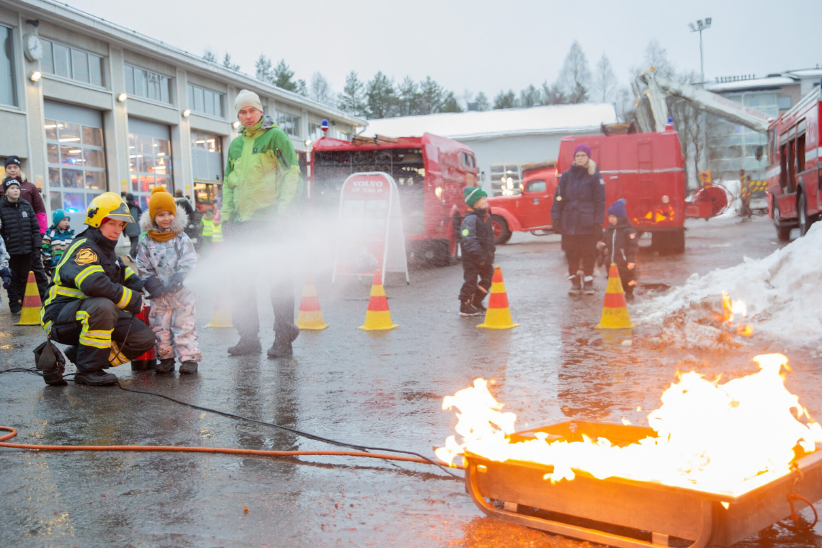 Pelastaja sytyttää rasvapaloa. Taustalla seisoo paljn lapsia ja aikuisia seuraamassa sammutusnäytöstä.