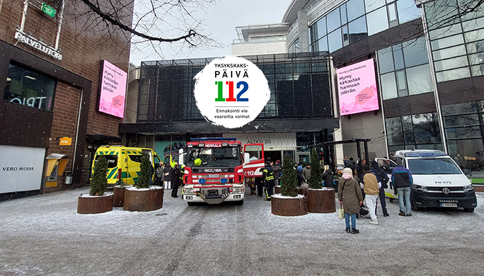 Kauppakeskuksen edustalla ihmisiä tutustumassa ambulanssiin, paloautoon ja poliisiautoon. Kuvassa myös 112-päivän logo.