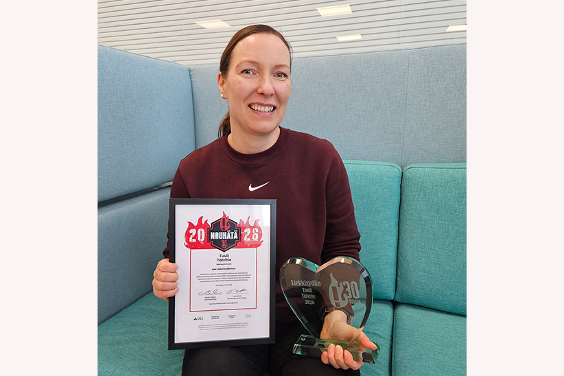 Henkilö istuu vaaleansinisellä sohvalla sisätiloissa ja pitää kädessään kehystettyä kunniakirjaa sekä sydämenmuotoista palkintopokaalia; taustalla on vaaleita seiniä ja toimistomainen ympäristö.