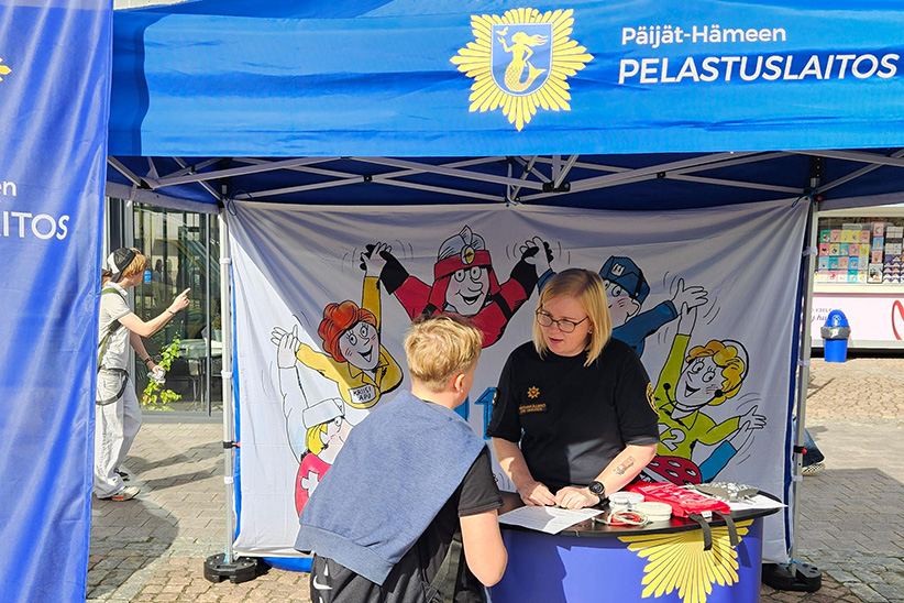 Pelastuslaitoksen sinisen teltan edessä järjestetyssä tapahtumapisteessä henkilö neuvoo lasta pöydän äärellä. Taustalla näkyy sarjakuvamaisia pelastushahmoja esittävä kangas sekä pelastuslaitoksen tunnukset.