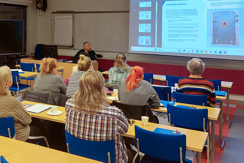 Luokkahuoneessa pidettävä koulutustilaisuus, jossa aikuiset osallistujat istuvat pulpeteissa ja kuuntelevat kouluttajaa. Valkokankaalle on heijastettu esitysmateriaalia.