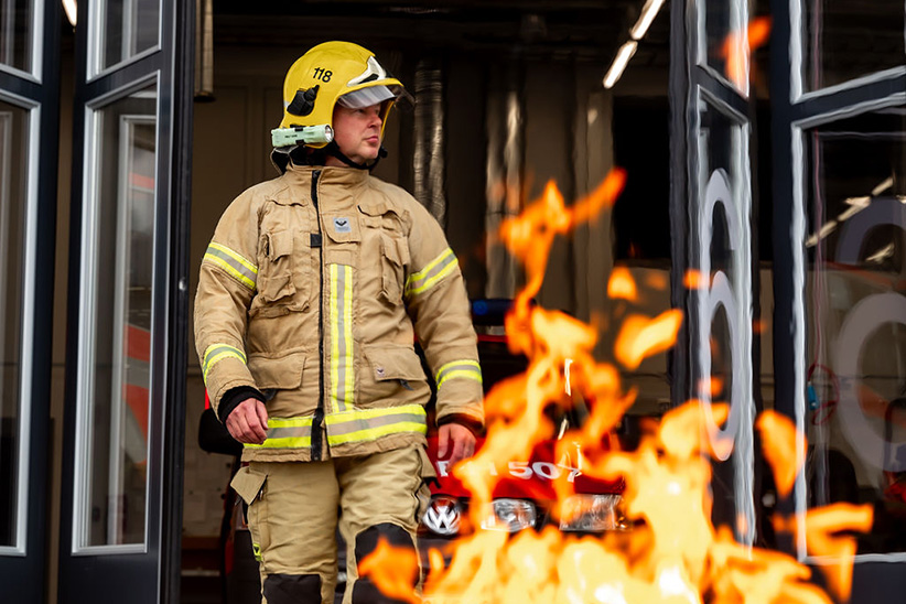 Pelastaja sammutusvarusteissa kypärä päässän kävelee ulos paloauton tallista ja edessä liekkejä.