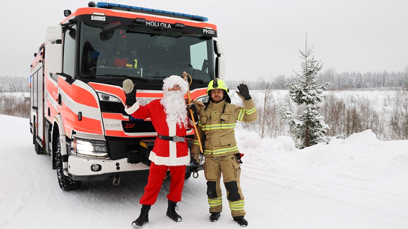 Pelastaja ja joulupukki vilkuttavat lumisessa maisemassa. Takana paloauto.