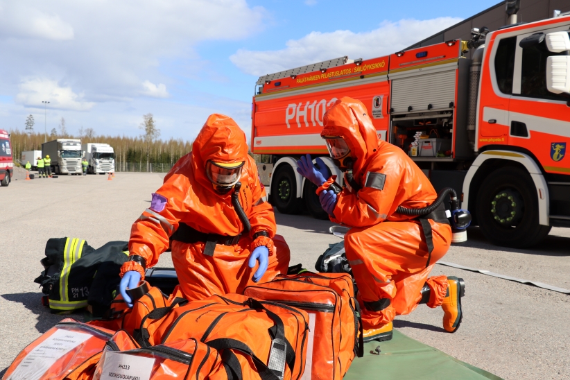 Oranssiin suoja-asuun pukeutuneita henkilöitä avaamassa laukkuja, taustalla paloauto.