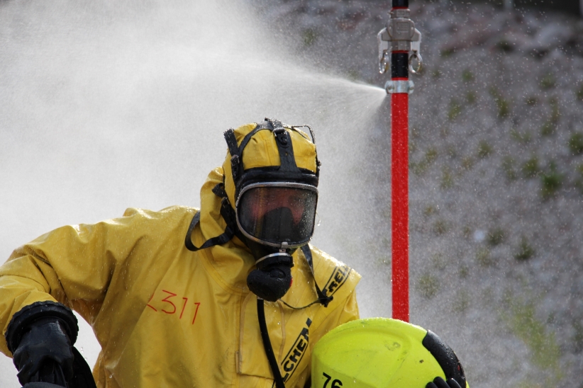 Keltaiseen suoja-asuun pukeutunut henkilö kaasunaamari kasvoilla vesisuihkussa.