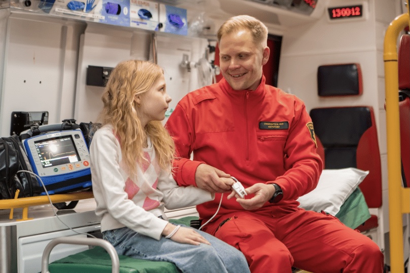 Hymyilevä ensihoitaja ja lapsipotilas istuvat ambulanssin hoitotilassa. Ensihoitaja kiinnittää mittaria lapsen sormeen. 