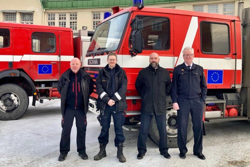 Kaksi punaista paloautoa Tampereen keskuspaloaseman pihassa. Ajoneuvojen kyljissä EU:n lipputarrat. Ajoneuvojen edessä seisoo neljä mieshenkilöä virkavaatteissaan.