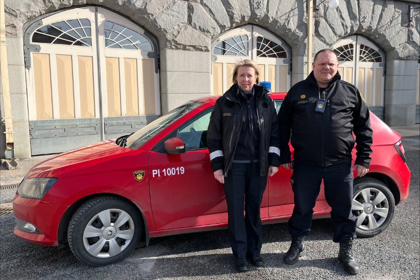 Kaksi palotarkastajaa pelastuslaitoksen virkavaatteissa Keskuspaloaseman pihalla. Taustalla punainen virka-auto.