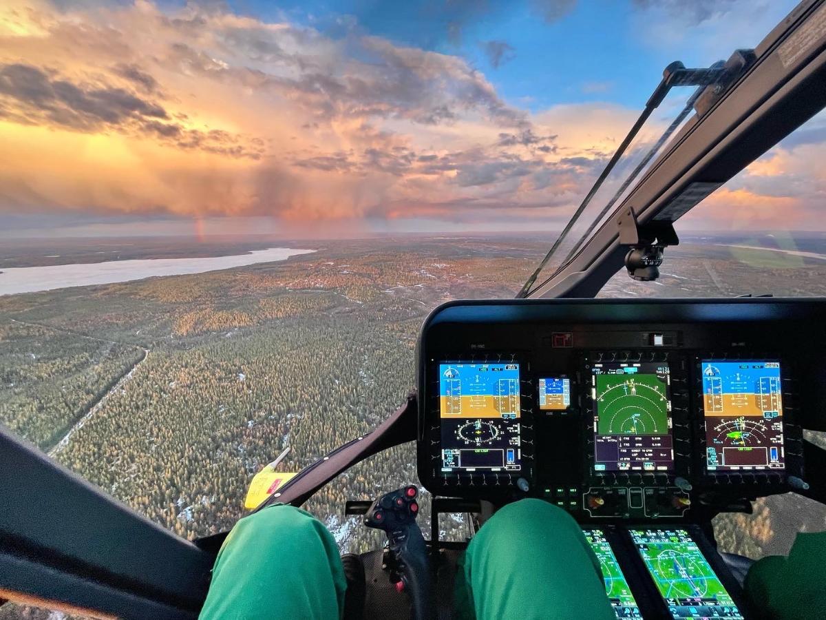 Lentäjä istuu lentävässä helikopterissa, alhaalla talvinen metsämaisema