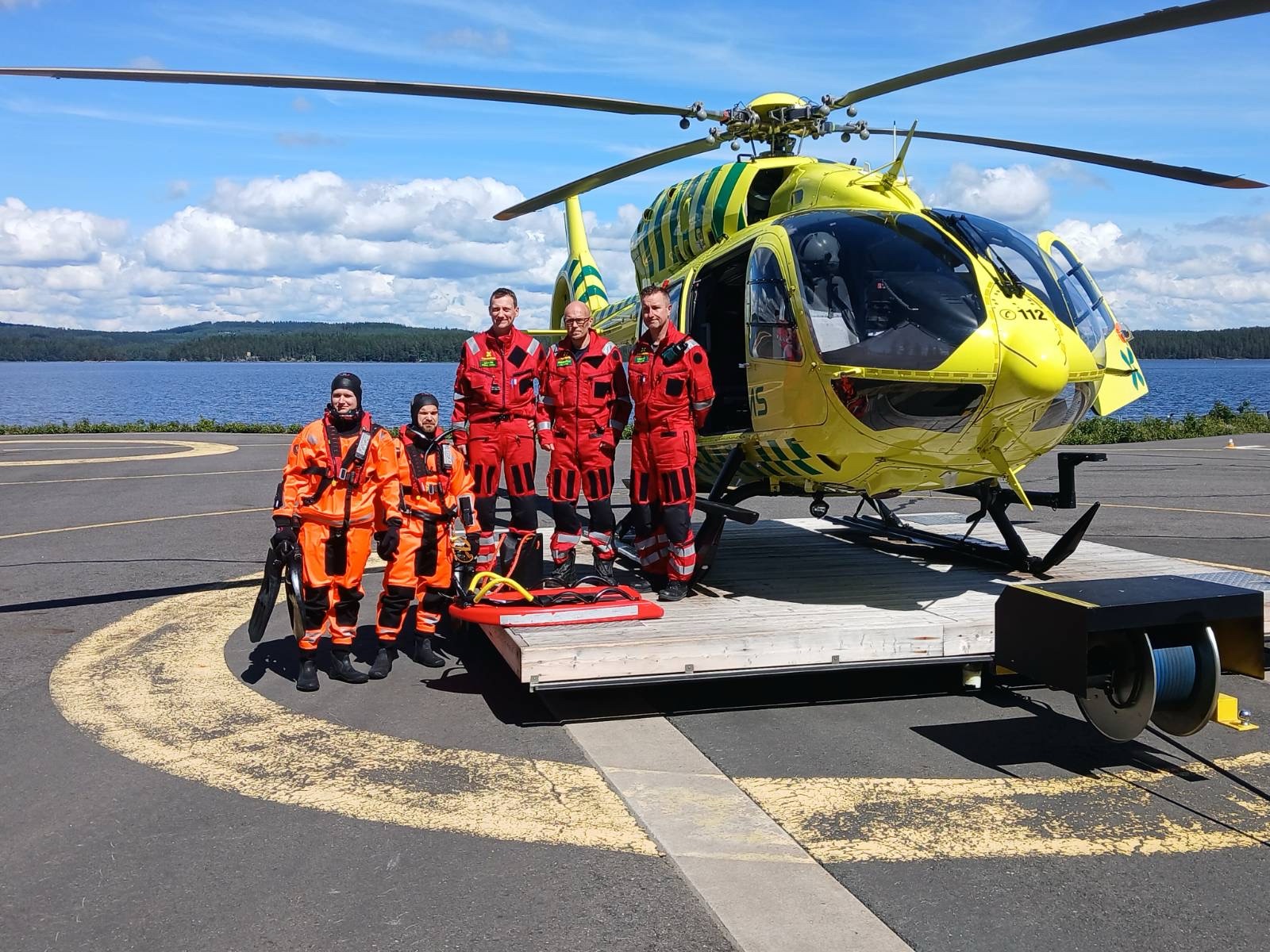 Helikopterin edessä kolme punaisiin pukeutunutta ensihoidon henkilökuntaa sekä kaksi pelastajaa pintapelastuspuvuissa.