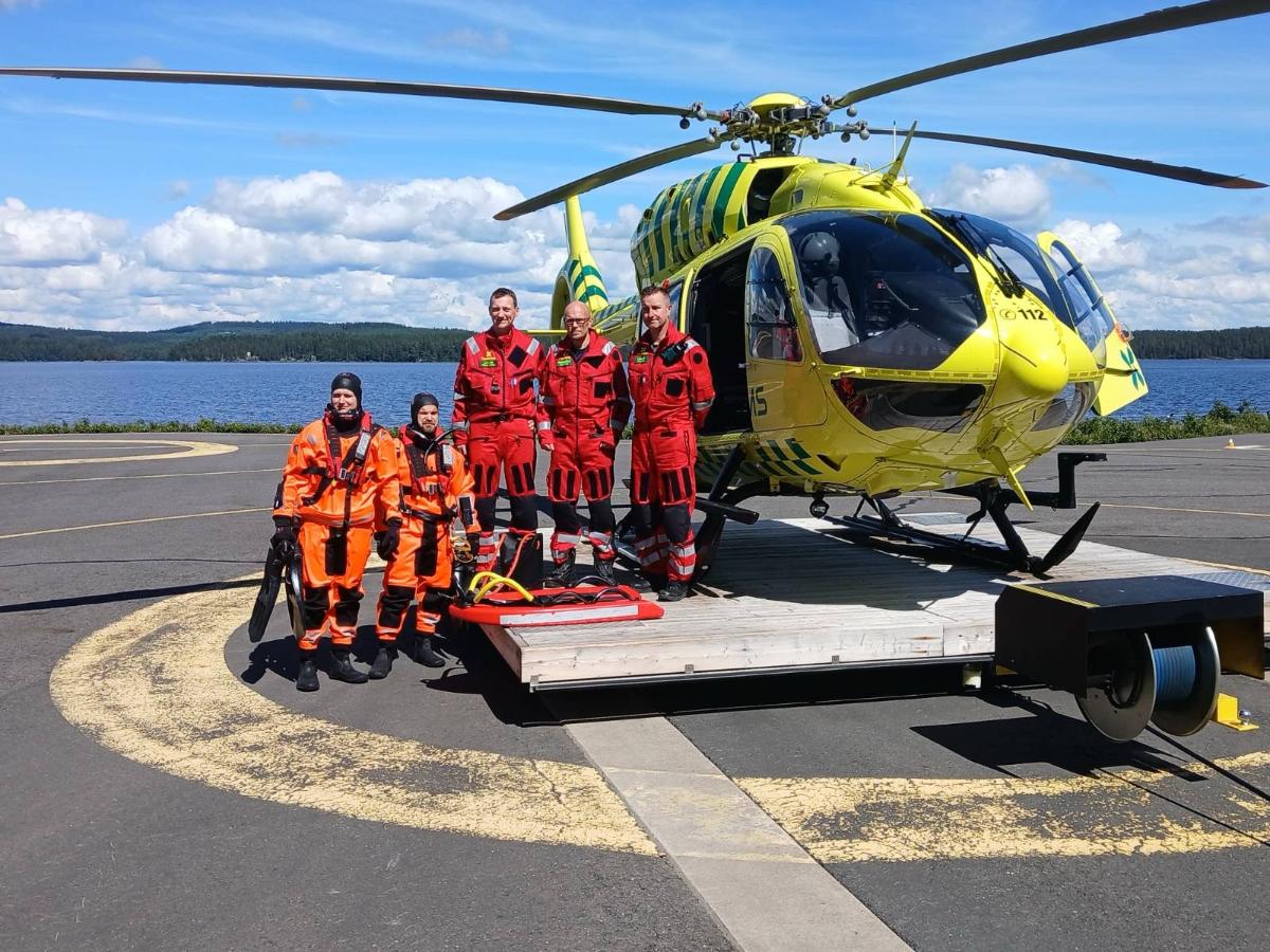Helikopterin edessä kolme punaisiin pukeutunutta ensihoidon henkilökuntaa sekä kaksi pelastajaa pintapelastuspuvuissa.