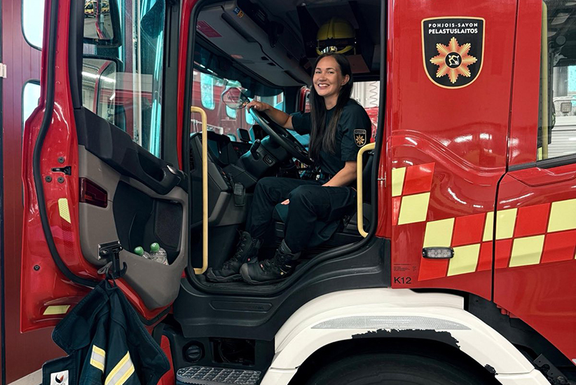 Merika Rasila istuu sammutusauton ohjaamossa ja hymyilee leveästi kameralle. Auton ovi on auki ja siitä roikkuu pelastusasun takki.
