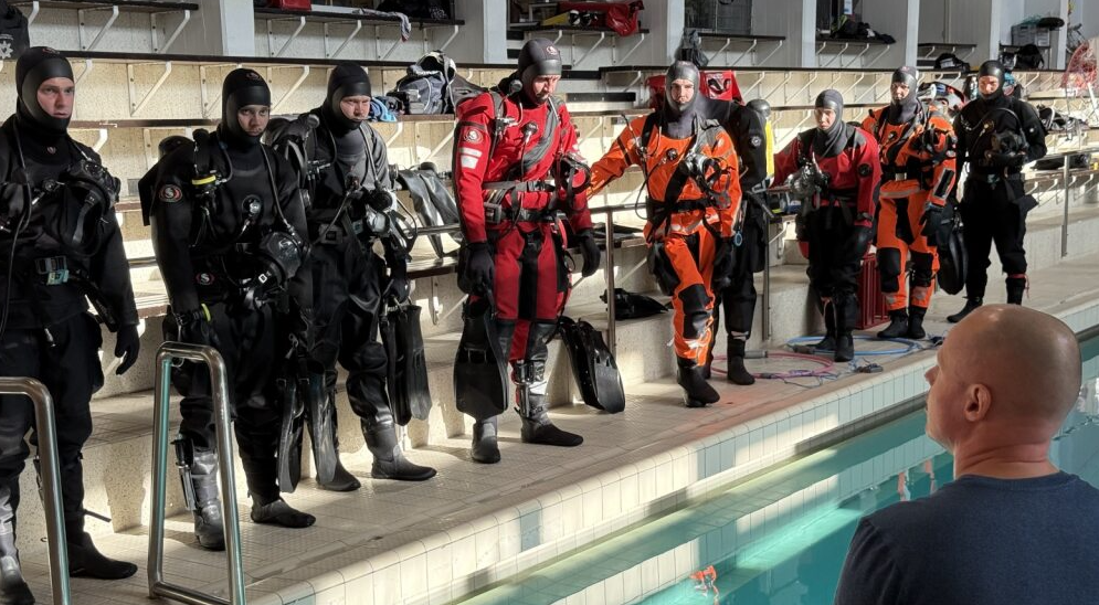 Sukeltajat seisovat altaan reunalla ja kuuntelevat ohjaajan ohjeita.