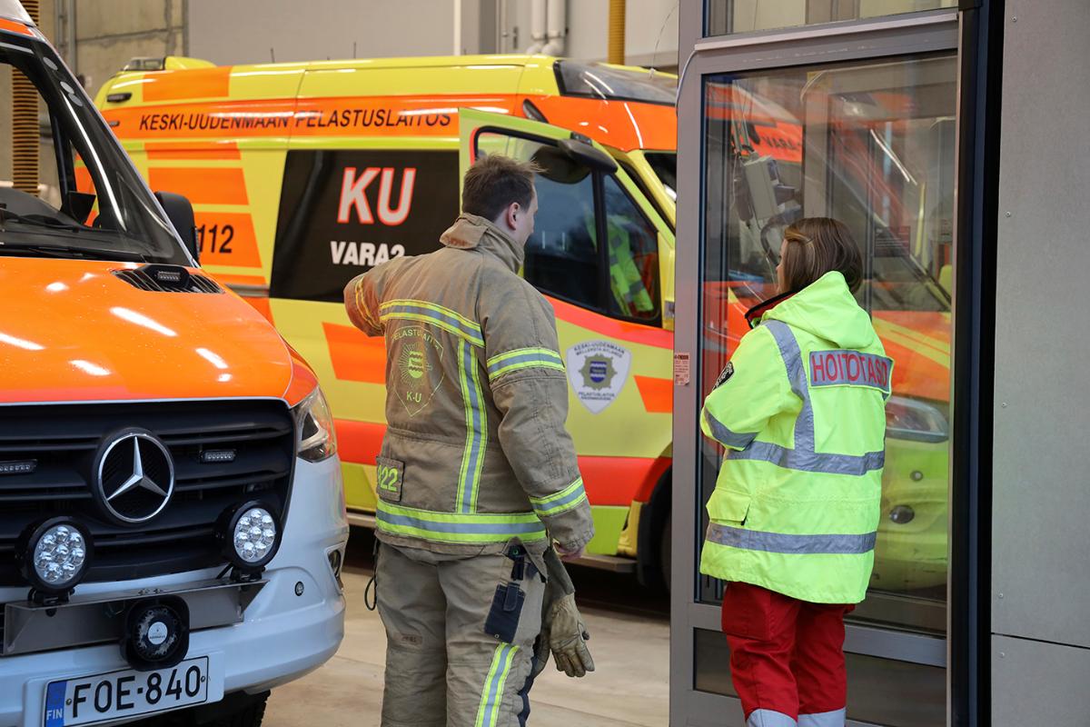 Palomies ja ensihoitaja keskustelevat selin. Taustalla kärkiyksikkö ja ensihoitoyksikkö.