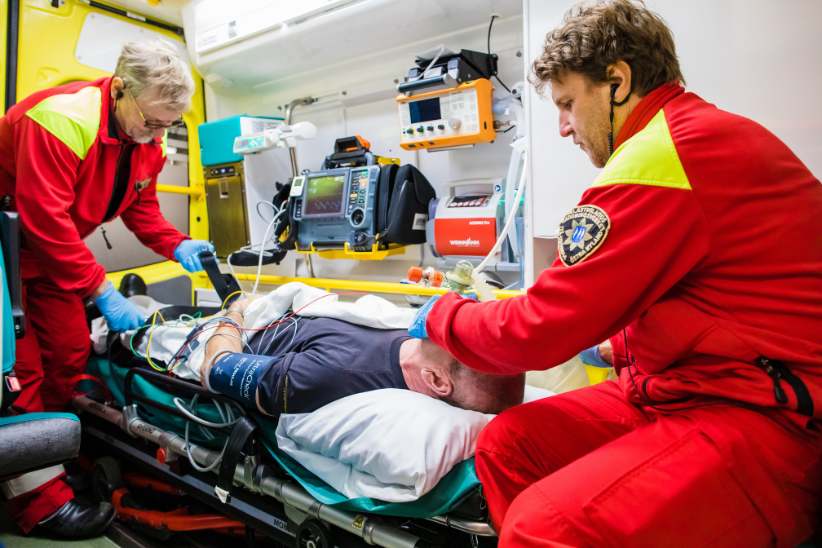 Kaksi ensihoitajaa hoitaa potilasta ambulanssissa. Potilas makaa paareilla. Potilaalle on laitettu happimaski.