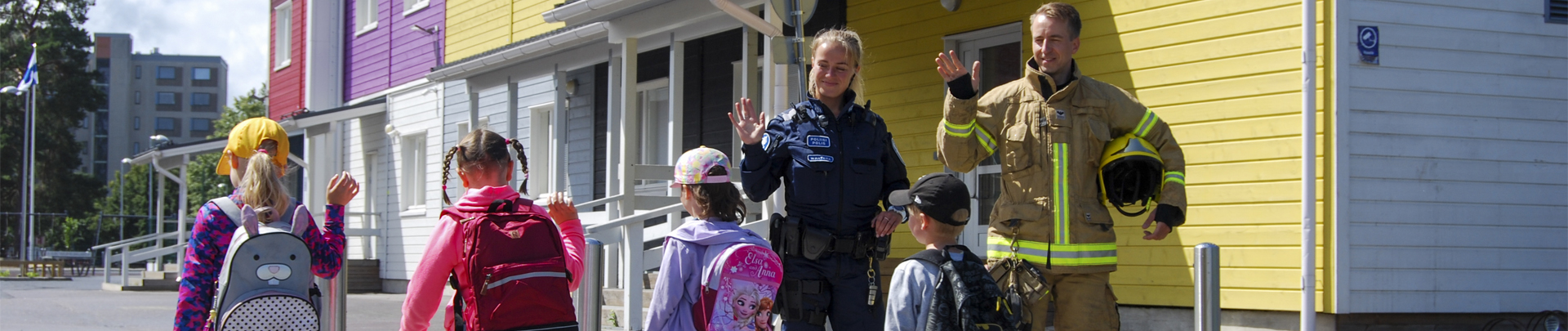 Palomies ja poliisi tervehtivät pieniä koululaisia, jotka ylittävät suojatien.
