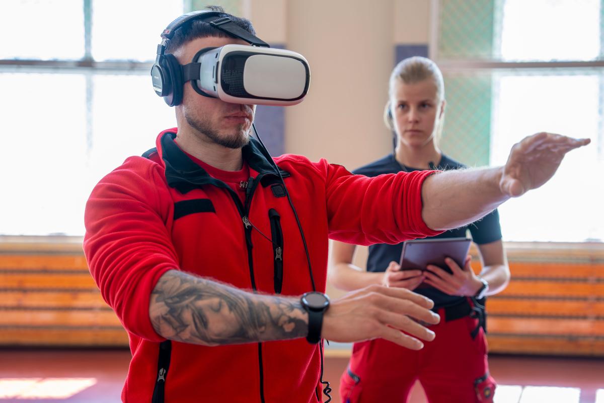 Miesensihoitaja harjoittelee virtuaalitodellisuudessa, VR-lasit päässään. Ensihoitajanainen ohjaa ja merkkaa asioita ylös tablettitietokoneelle.