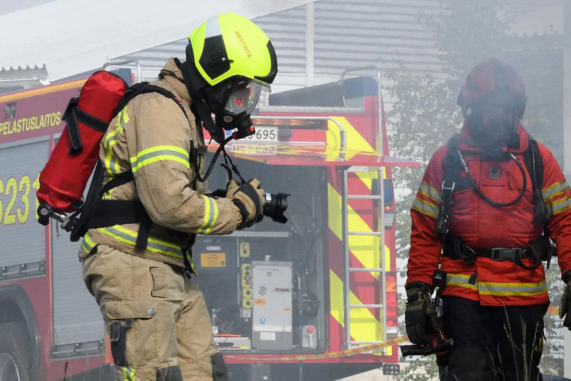 Pelastajan sammutusasussa oleva mies kuvaa kameralla palavaa merikonttia. Taustalla näkyy pelastusryhmänjohtaja punaisessa sammutusasussaan.