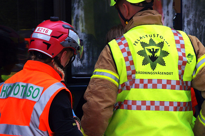 Ensihoitaja ja pelastustyöntekijä selkäpuolelta niin, että heidän varusteidensa selkämykset näkyvät.