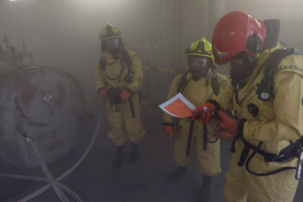 Kolme keltaisiin suojapukuihin pukeutunutta pelastajaa savuisessa tilassa. Yhdellä on papereita käsissään.